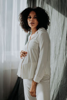 A pregnant woman wearing a white maternity pyjama set with straight-cut trousers and a top with side-seams and a collared neckline.