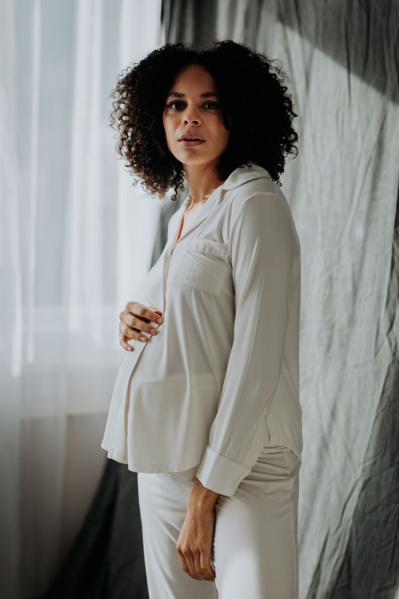 A pregnant woman wearing a white maternity pyjama set with straight-cut trousers and a top with side-seams and a collared neckline.